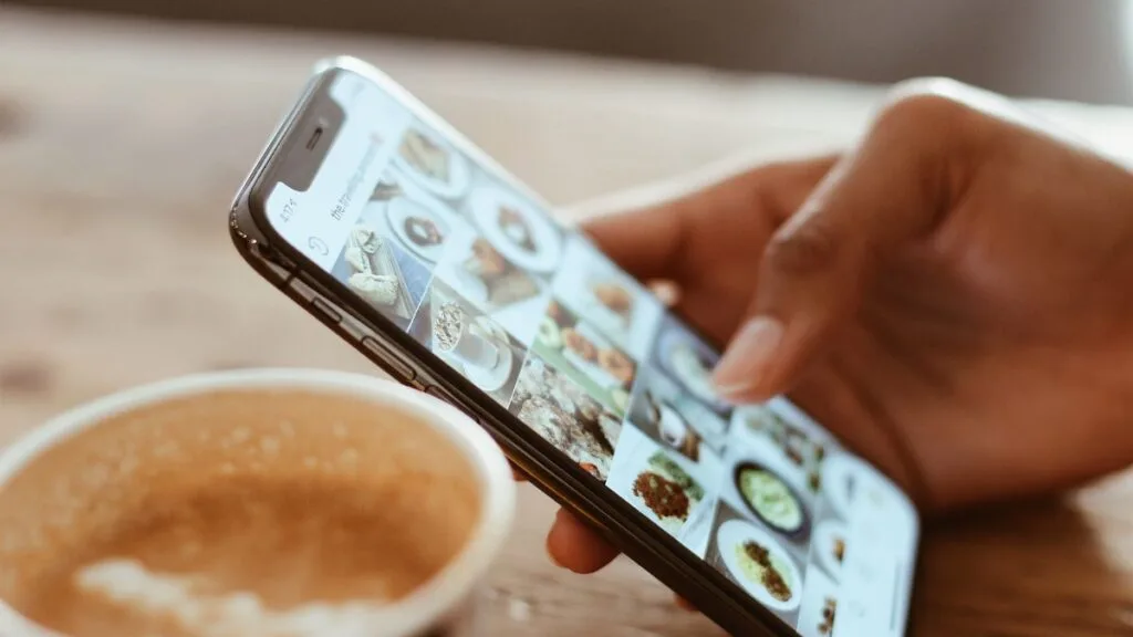 Person holding smartphone showing food recipe gallery while enjoying warm coffee cup in cozy setting