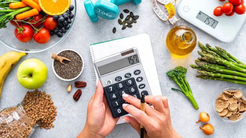 Hands using calculator surrounded by colorful fruits, vegetables, nuts, seeds, scale and measuring tape on marble surface
