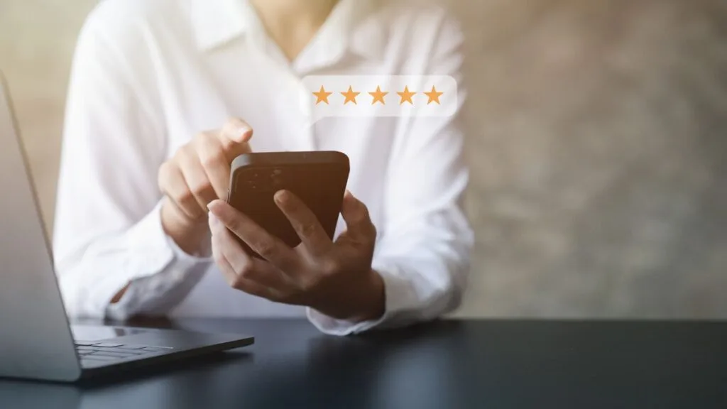 Person in white shirt holding smartphone showing five gold stars rating while sitting at desk with laptop