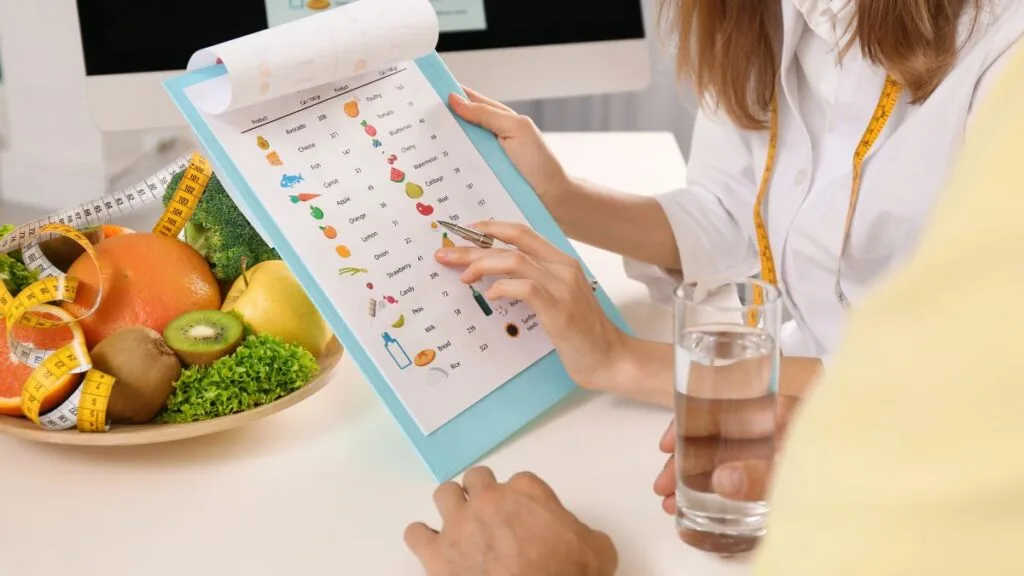 Female nutritionist in white coat reviewing colorful food chart with fresh fruits, measuring tape, and water glass on desk