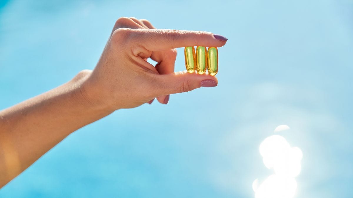 Woman's hand holding three golden vitamin capsules against bright blue sky with sunlight