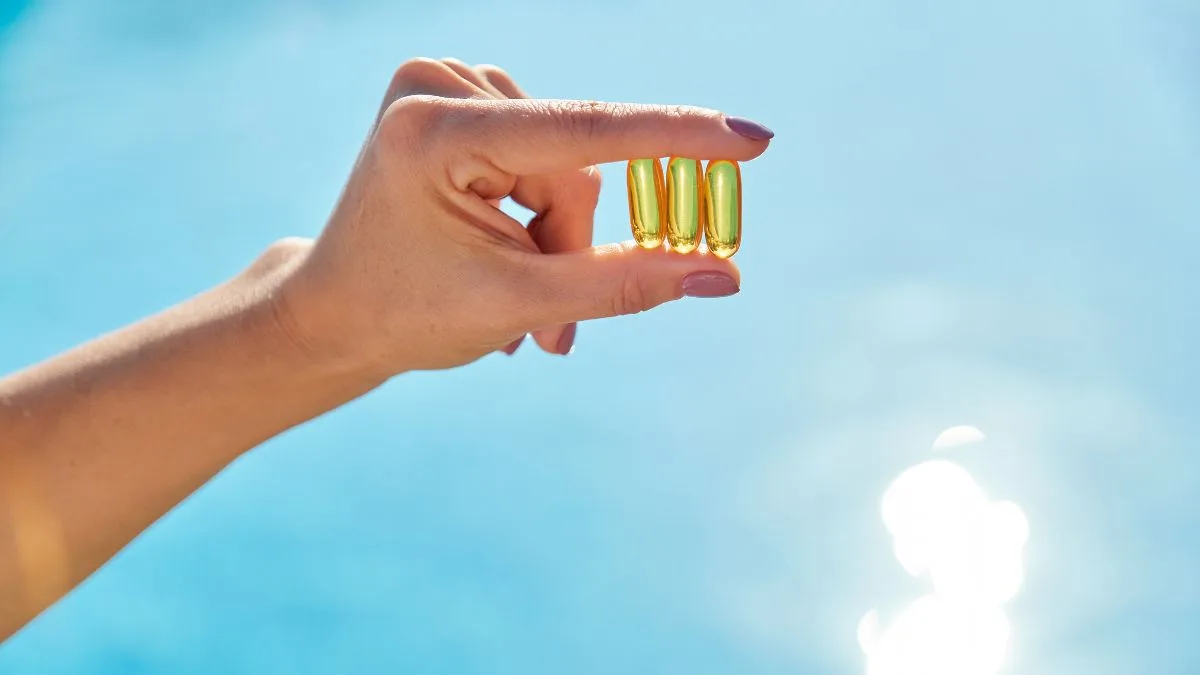 Woman's hand holding three golden vitamin capsules against bright blue sky with sunlight