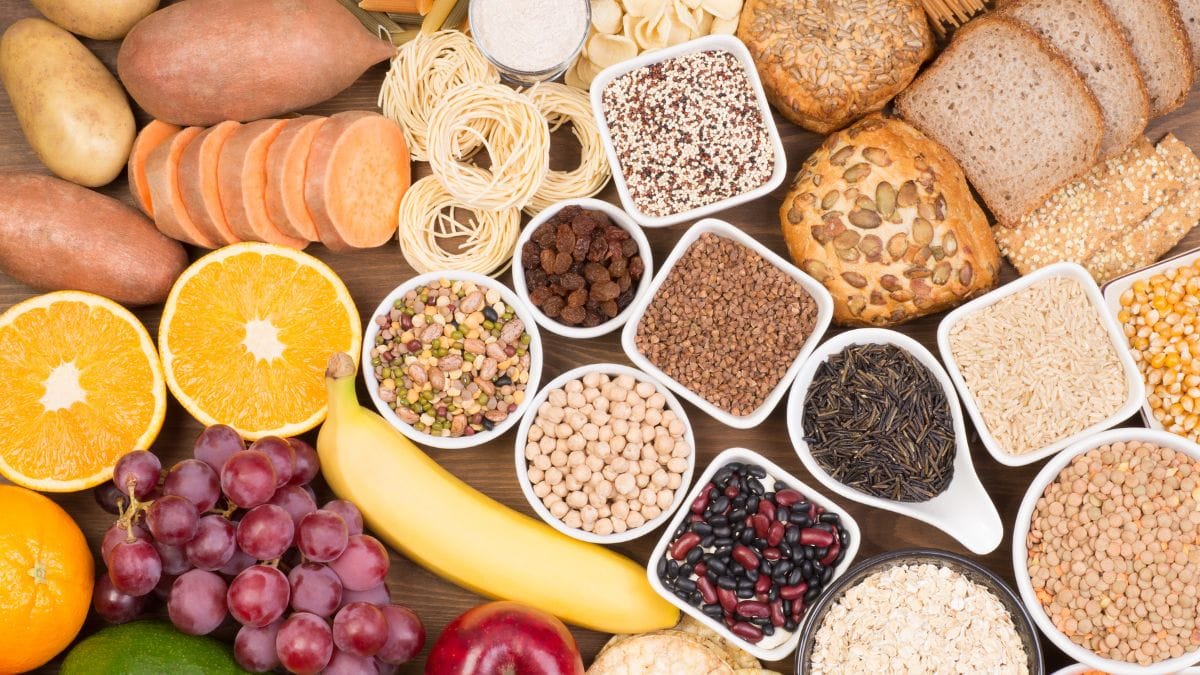 Wooden table with assorted high-fiber foods including whole grain bread, pasta, fruits and legumes in bowls