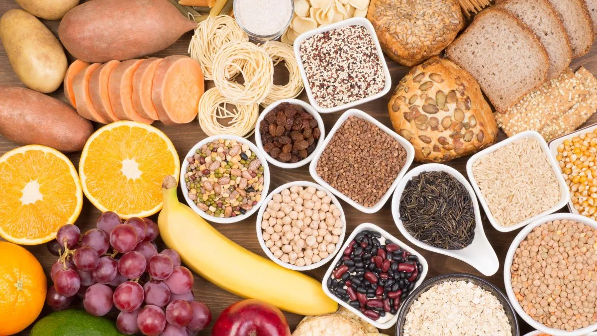 Wooden table with assorted high-fiber foods including whole grain bread, pasta, fruits and legumes in bowls