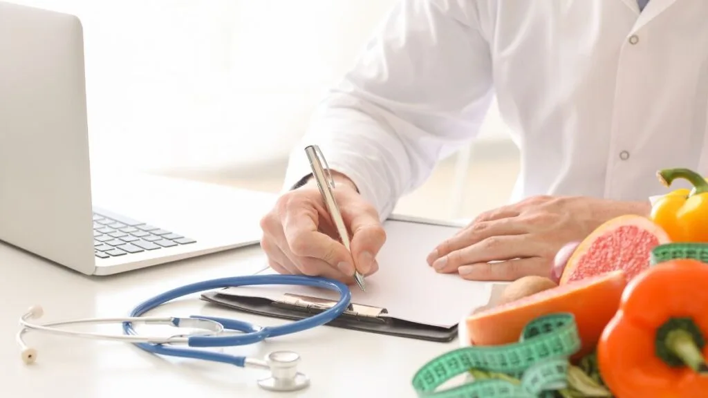 Healthcare professional in white coat writing nutrition plan at desk with stethoscope, laptop, and colorful fresh fruits