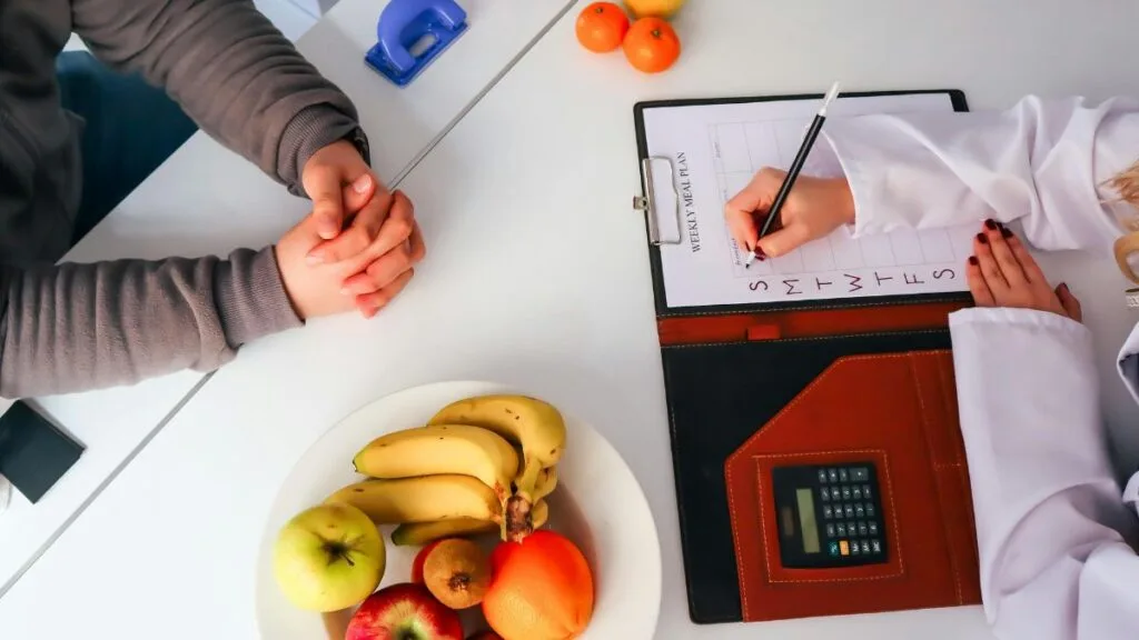 Nutrition counselor writing meal plan on clipboard while client sits across white desk with plate of fresh fruits