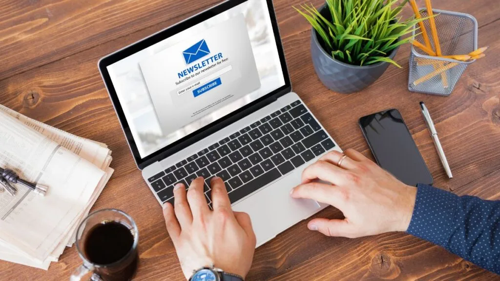 Hands typing on laptop showing newsletter subscription page on wooden desk with coffee, newspaper, phone, and plant