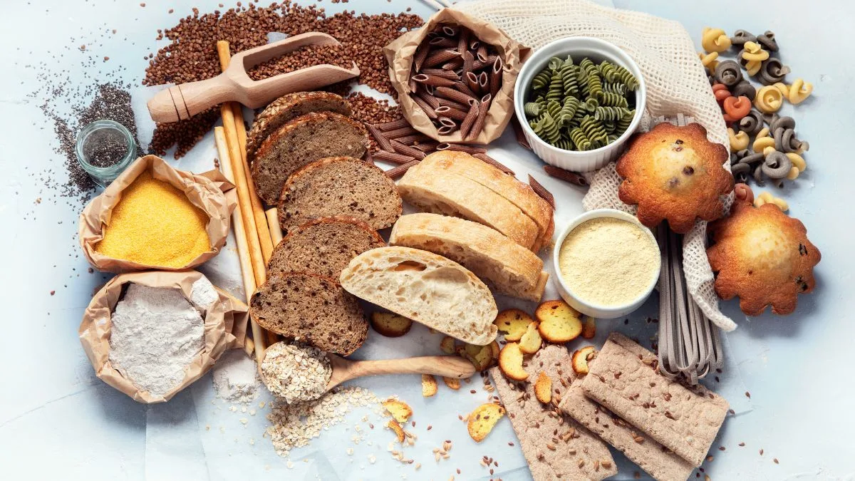Light blue surface with various whole grain products including sliced bread, pasta, flour and seeds