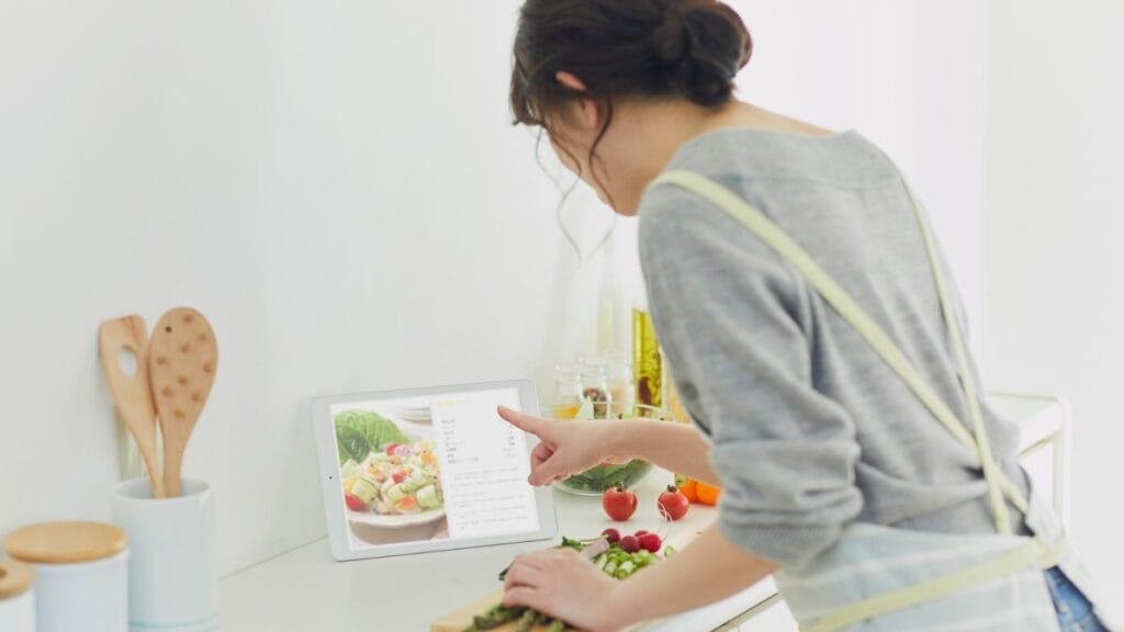 Woman in gray sweater preparing fresh vegetable salad while following cooking recipe on tablet in modern kitchen