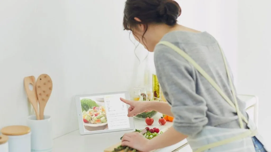Woman in gray sweater preparing fresh vegetable salad while following cooking recipe on tablet in modern kitchen